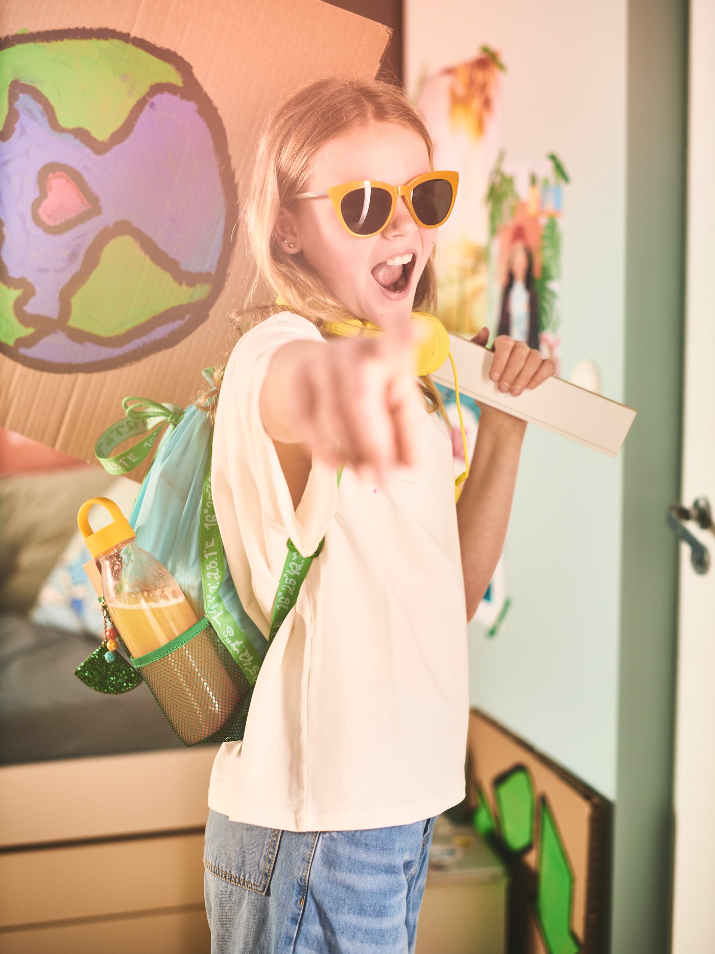 A girl, wearing sunglasses and a t-shirt, points forward, with BLÅVINGAD blue-green backpack on her back.