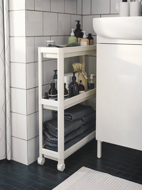 Un carrito blanco VESKEN ubicado en el espacio entre el mueble vanitorio y el muro del baño. En el carrito se ven toallas y productos de baño como jabones y recipientes varios.