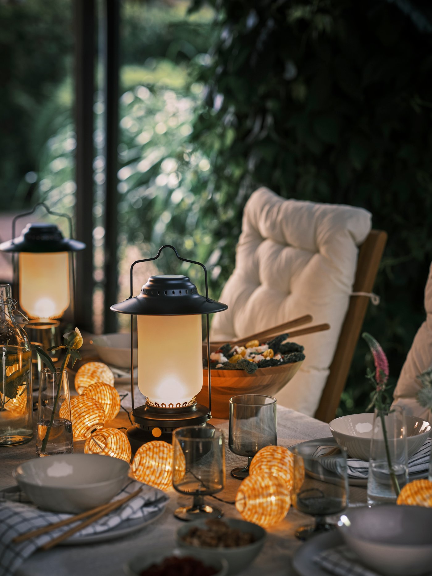 An outdoor table set with plates, bowls and glasses, plus two black STORHAGA table lamps and an LED lighting chain.