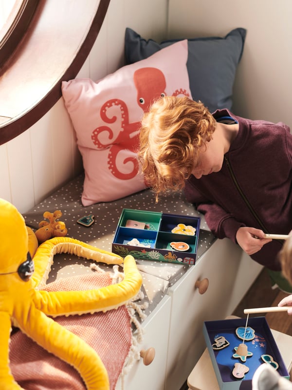 A boy plays with a BLÅVINGAD fishing game on a window seat with a cushion in a BLÅVINGAD octopus cushion cover and soft toy.