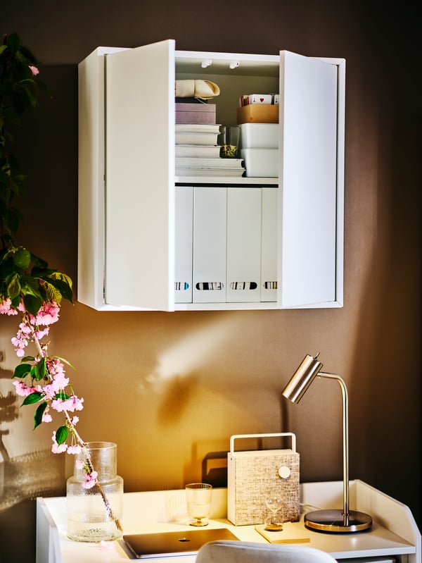 A white EKET cabinet with doors holding white KVISSLE magazine files hangs over a white HAUGA desk with a VIRRMO work lamp.