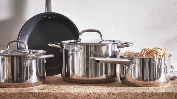 An IKEA cookware set, featuring stainless-steel pots, a frying pan, and a colander filled with dry pasta, is arranged on a cork countertop.