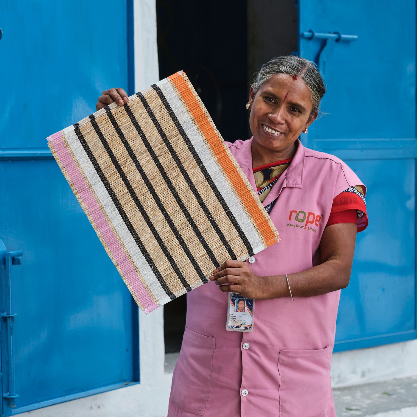 A person in pink clothes stands smiling in front of a pair of blue doors and holds up a striped MÄVINN place mat.