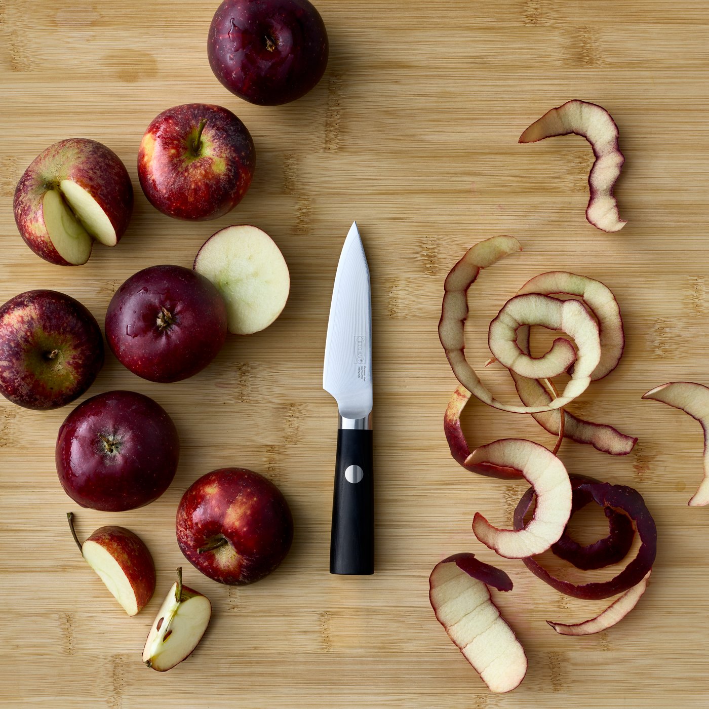 A close-up of a black FJÄLLRÖDING paring knife made of Damascus steel, surrounded by red apples and peels on a wooden surface.