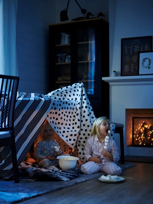 Two young girls eating marshmallows in a play campsite made of METTALISE and SKÄGGÖRT fabric draped over an EKEDALEN table.