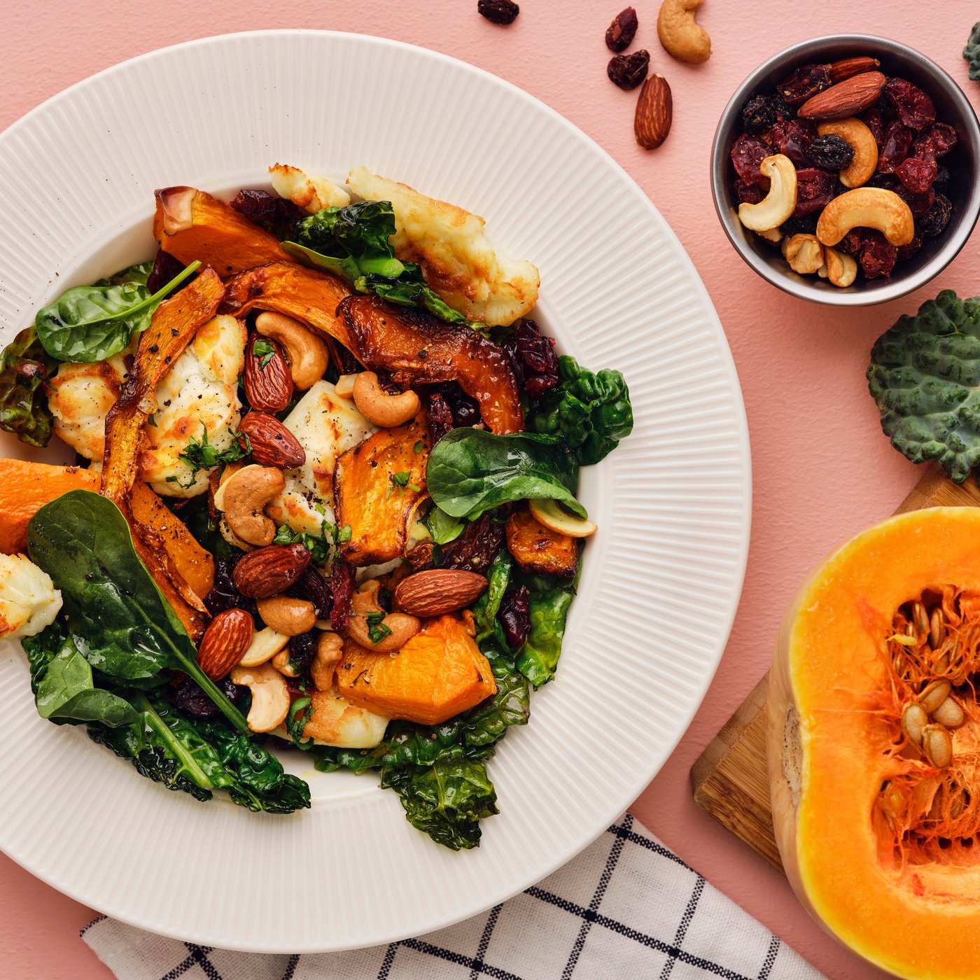 Roasted butternut squash, black kale and MUNSBIT roasted and lightly salted nuts and berries fill a dish on a pink tabletop.