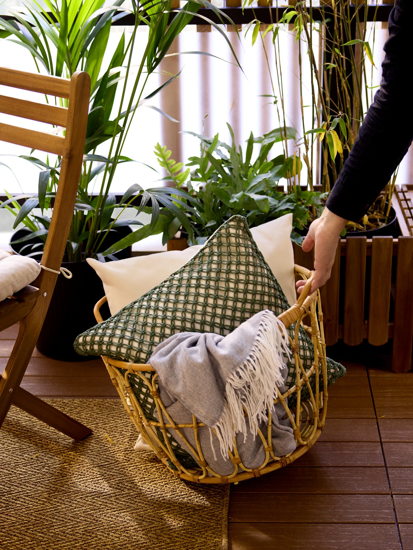 A hand is lifting a rattan SNIDAD basket by a handle. The basket holds cushions and throws that are temporarily on a balcony.