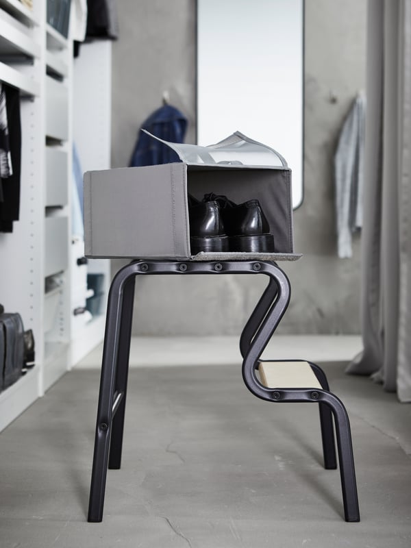 An open dark gray SKUBB shoe box containing a pair of black shoes placed on top of a black/birch GRUBBAN step stool.