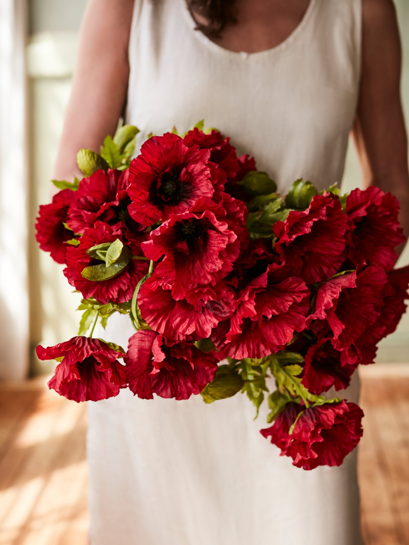 A big bunch of red ANLEDNING artificial poppy flowers with some green leaves held by a woman in front of her beige dress.