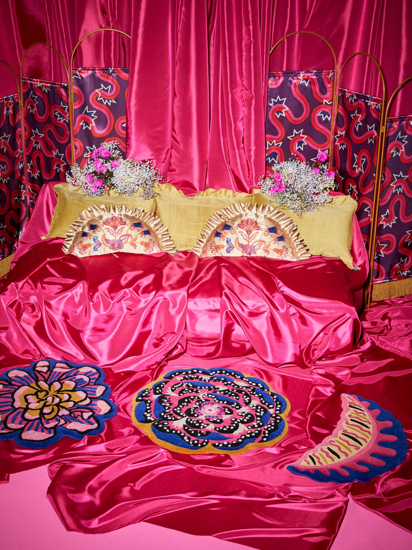Set of three KARISMATISK wool rugs in flower and leaf shapes are on a pink satin floor; gold cushions are in the background.