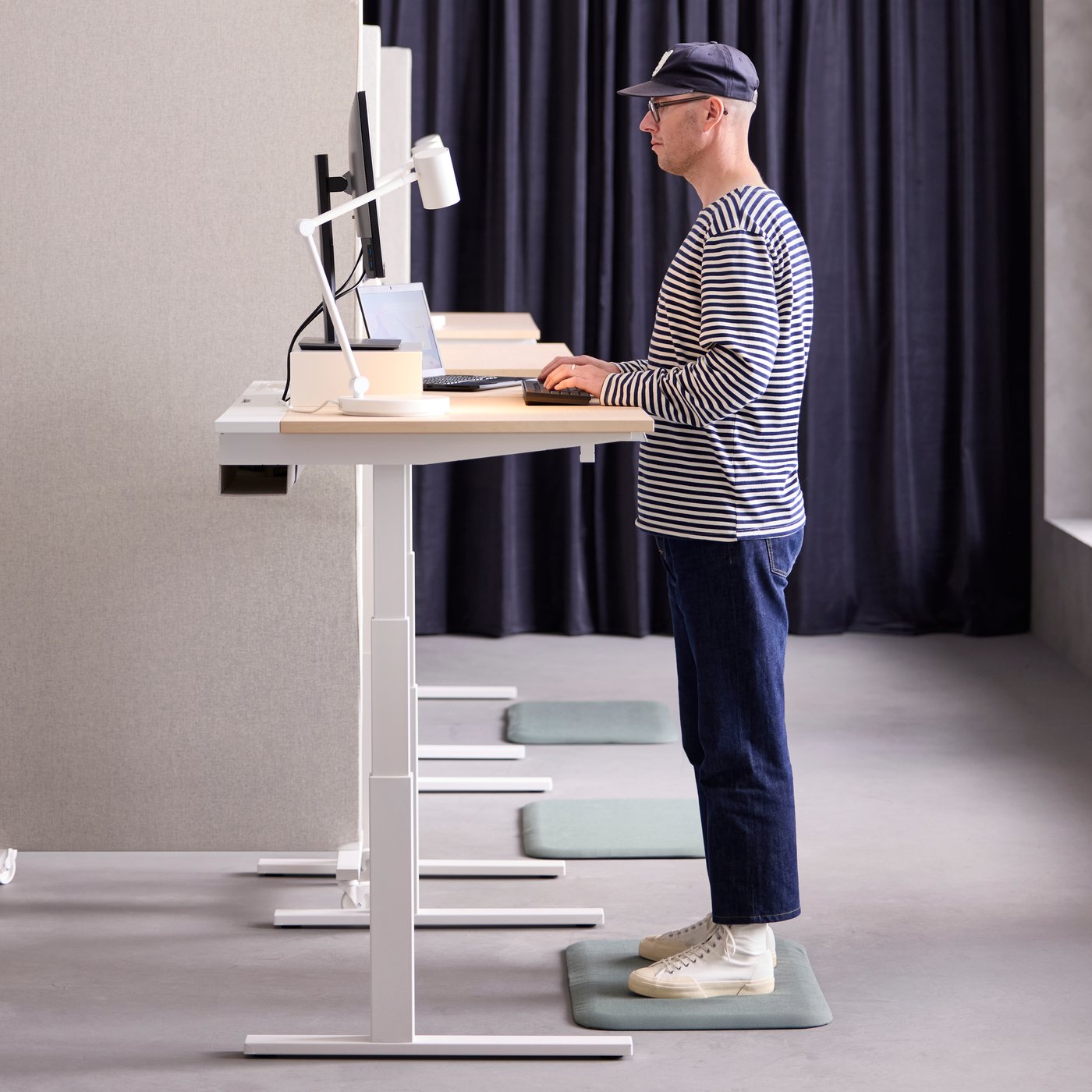 A person wearing a cap and glasses is standing behind a MITTZON sit/stand desk.