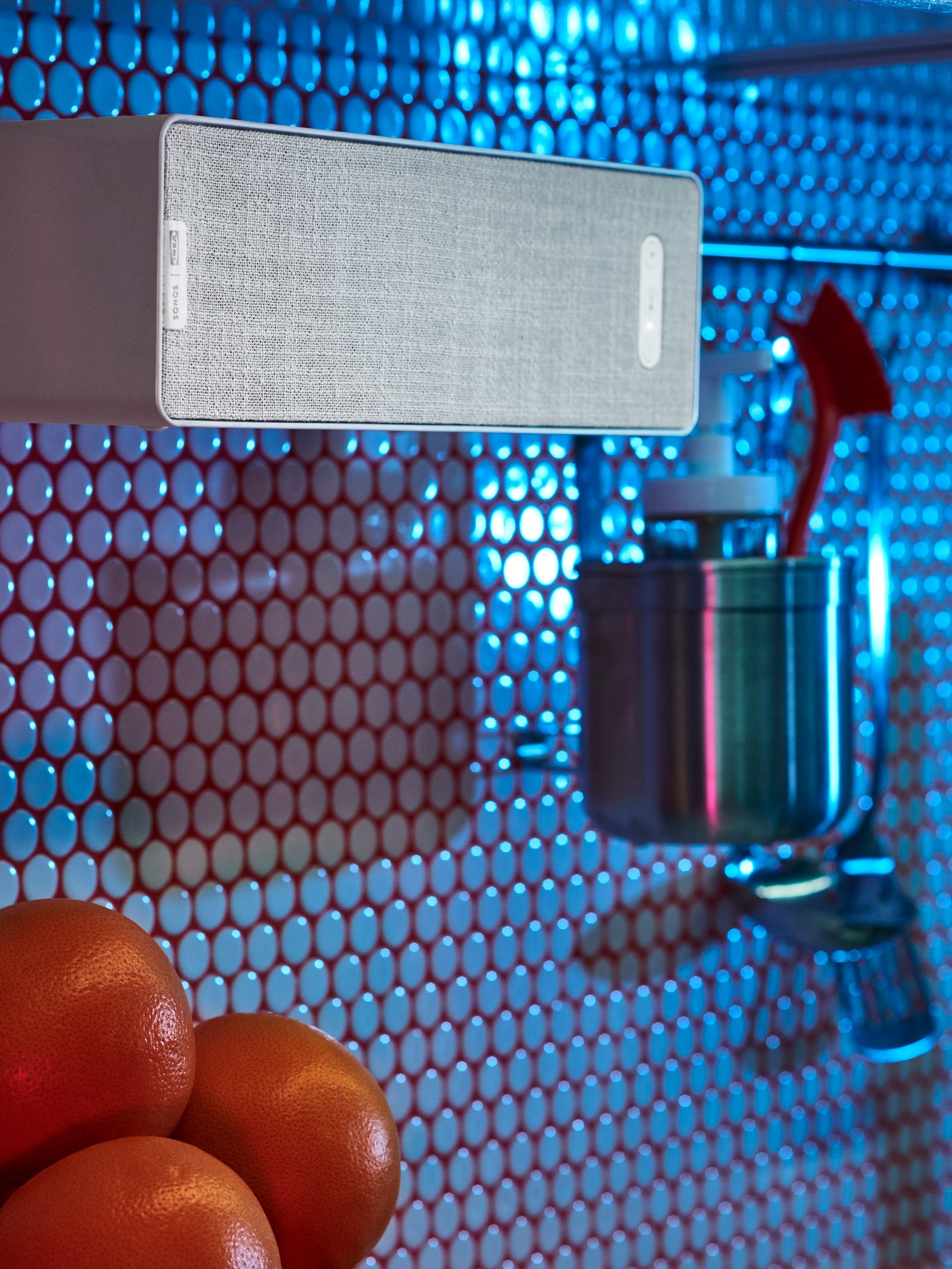 A white SYMFONISK WiFi bookshelf speaker hanging on a KUNGSFORS stainless steel rail on a kitchen wall with round tiles.