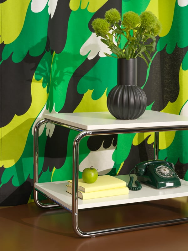 A white BAGGBODA side table stands in front of the ÄNGSFIBBLA pre-cut fabric. The table holds a black vase and an old phone.