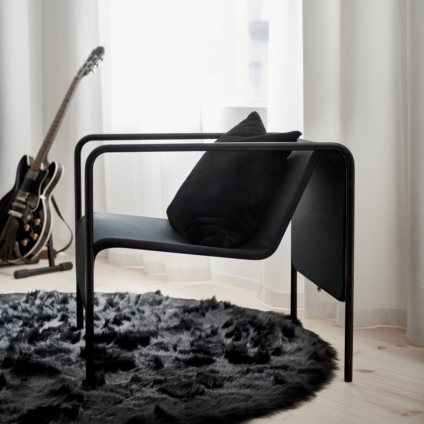 A black OBEGRÄNSAD armchair, rug and cushion cover on wooden floorboards next to an electric guitar and white curtains.