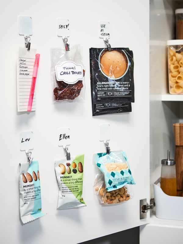 Die Innenseite einer Schranktür mit weissen selbst klebenden Kunststoffhaken und silberfarbenen Clips, an denen Tüten mit Snacks und Sossenpulver hängen