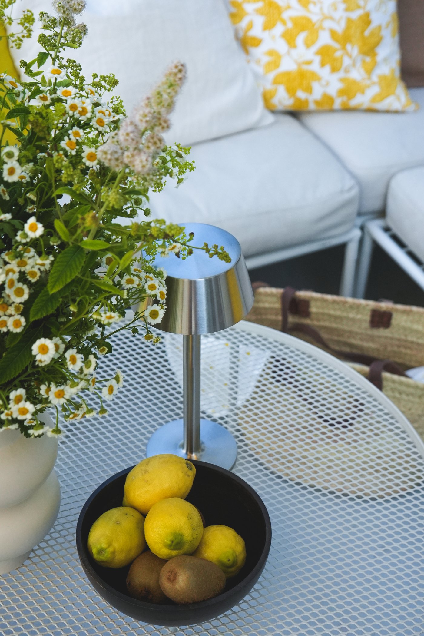 Weisser KROKHOLMEN Metall-Gartentisch mit silberner Lampe, schwarzer Schale mit Zitronen/Kiwis, weisser Vase mit Blumen. Im Hintergrund weisses Sofa mit gelbem Kissen.