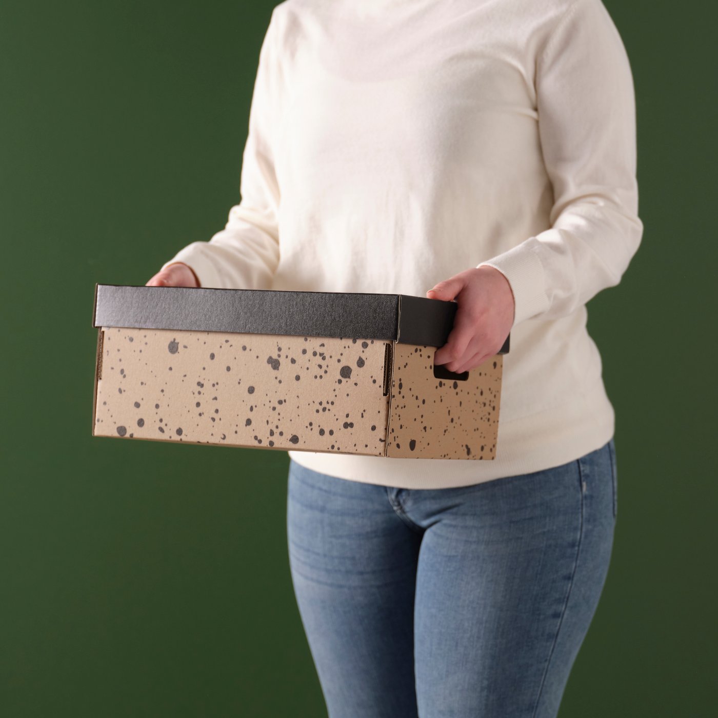 Eine Person trägt eine rechteckige Aufbewahrungsbox in den Händen. Die Box ist beige mit einem dezenten schwarzen Punktmuster und einem dunkelgrauen Deckel.