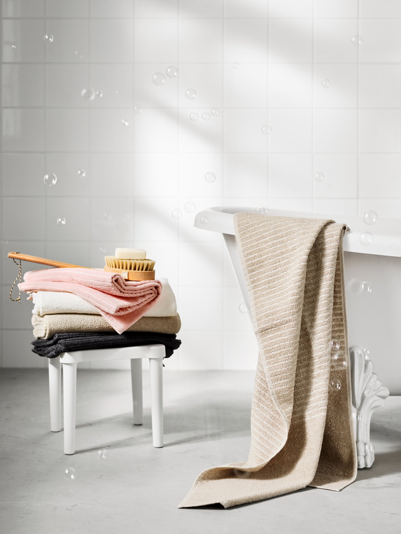 A pile of VÅGSJÖN bath towels in pink, white, light beige and dark grey on top of an UTTER stool at the end of a bath.