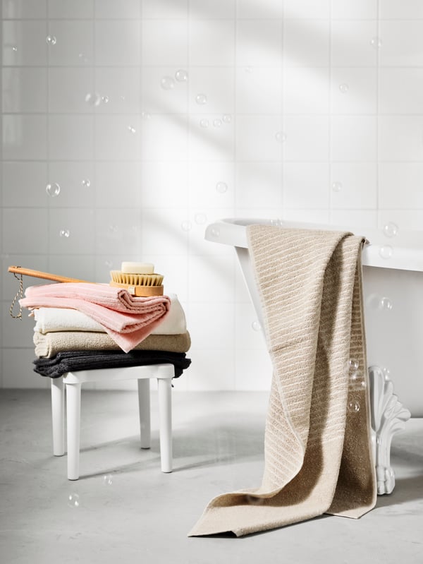 A pile of VÅGSJÖN bath towels in pink, white, light beige and dark grey on top of an UTTER stool at the end of a bath.