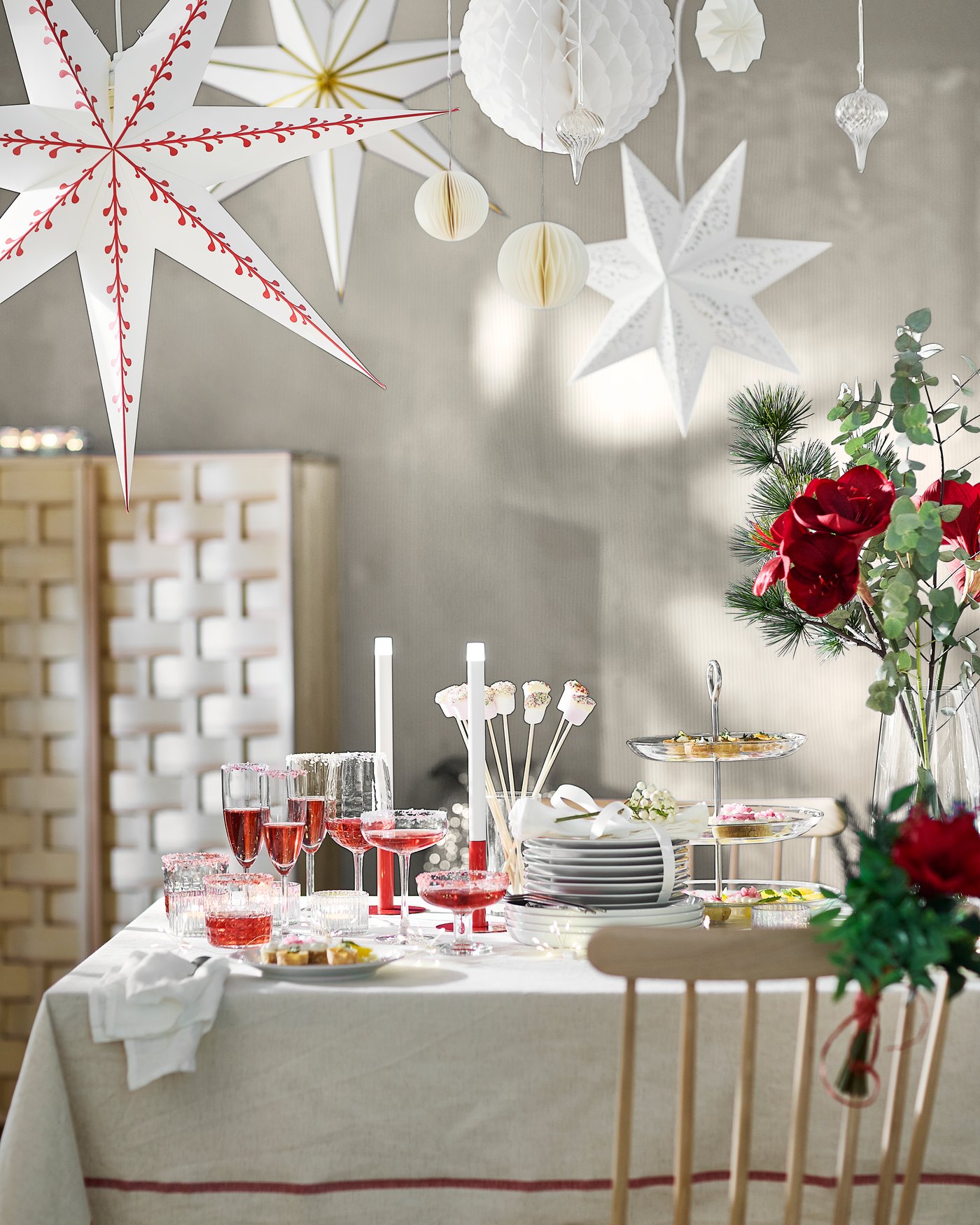 Christmas table set with tableware, glasses and candles, decorated with red flowers and large hanging paper stars.
