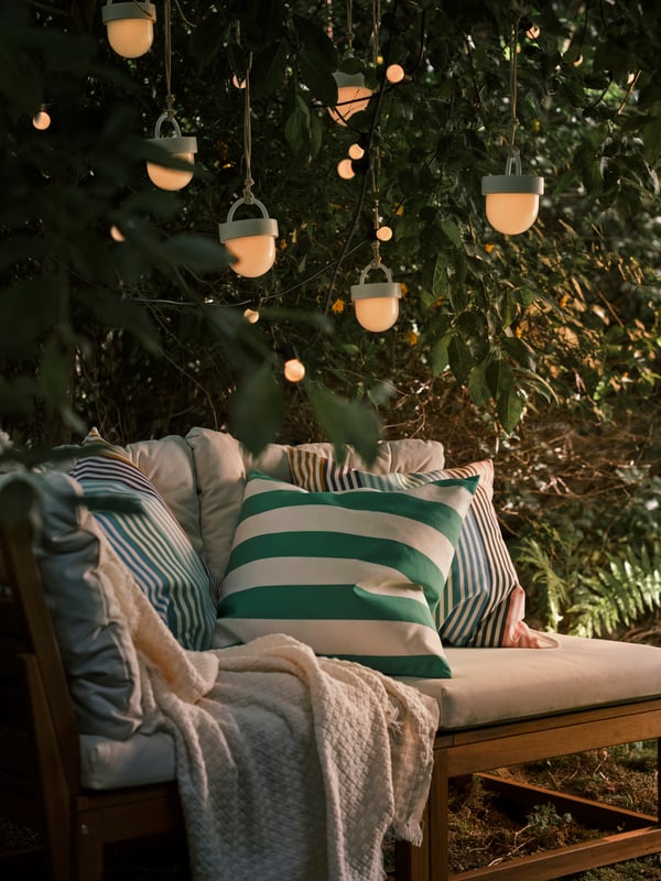 Several white SOMMARLÅNKE pendant lamps and a black lighting chain hanging in a tree above a light brown outdoor sofa.