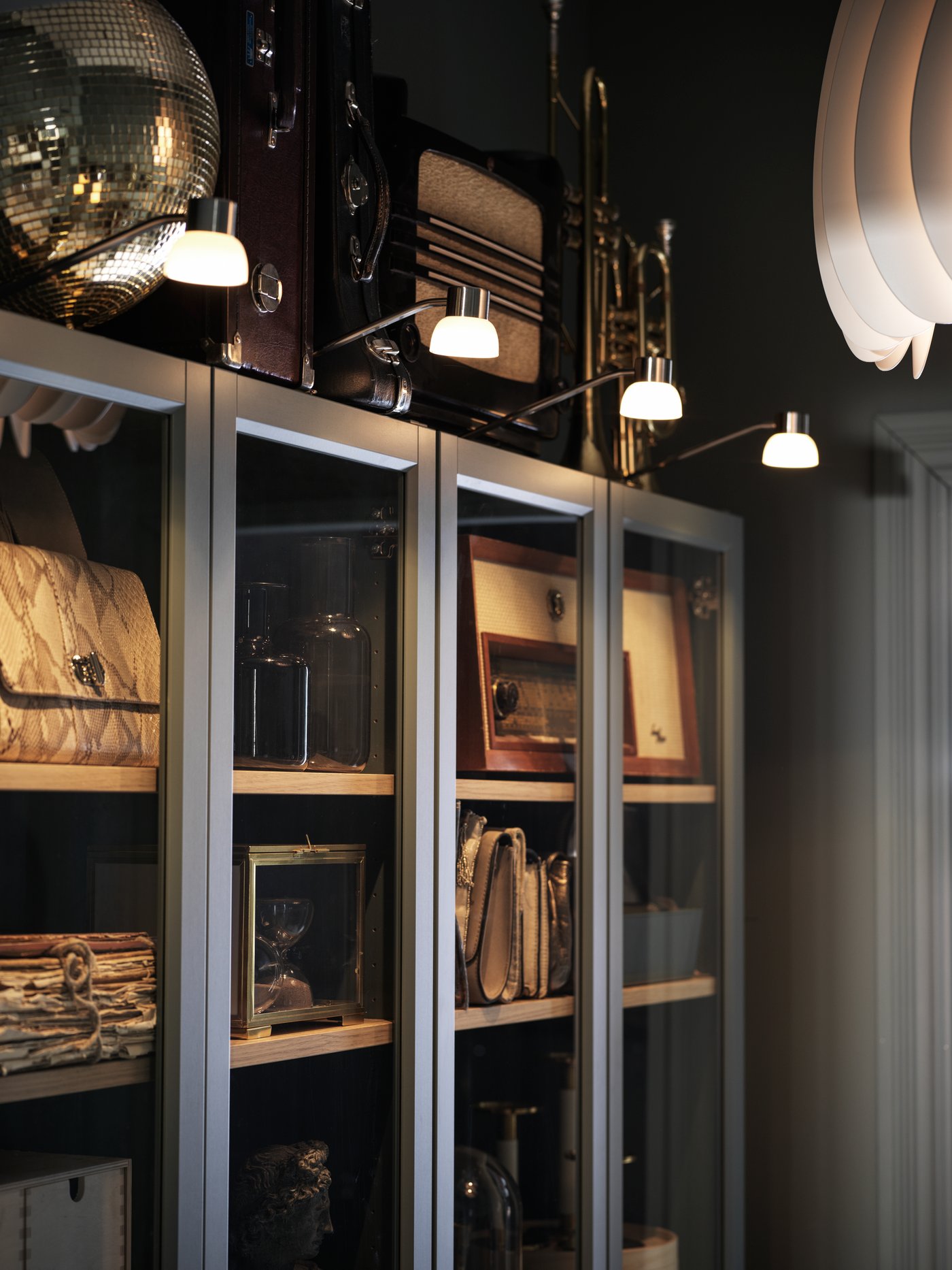 Storage in and on top of grey-turquoise BILLY glass-door bookcases. LINDSHULT LED cabinet lighting lights up their fronts.