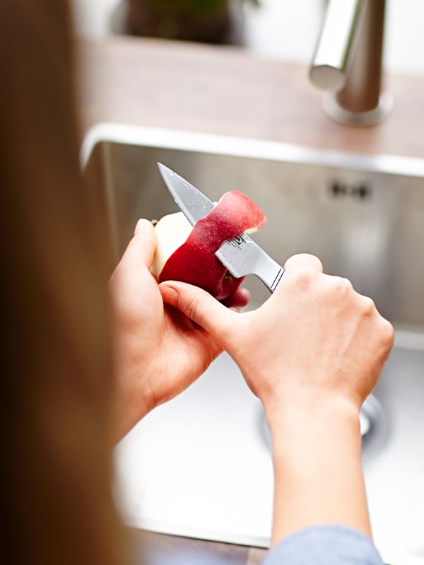 Hands are pealing a red apple with a paring knife in stainless steel.