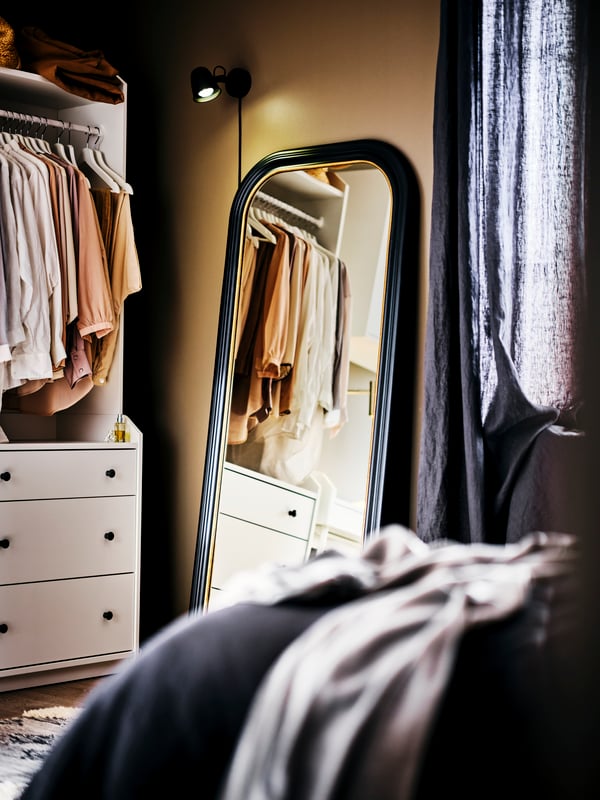 SKURUP wall lamp is mounted above the ALMAROD mirror next to the HAUGA open wardrobe in the bedroom.