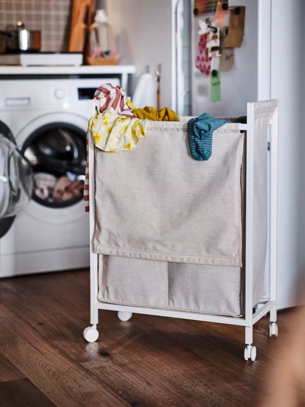 Clothes hanging over the edge of a white ENHET laundry bag with castors in front of a washing machine inside a white kitchen.