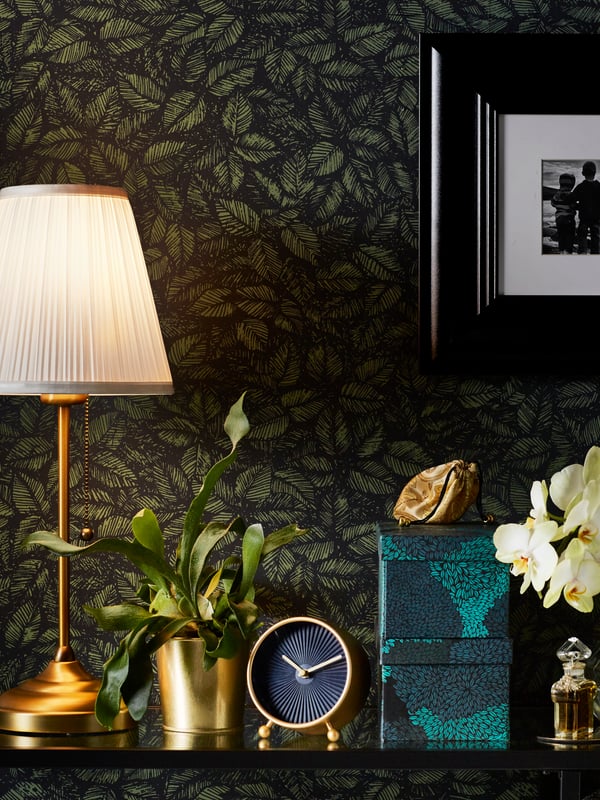 Une lampe de table ÅRSTID, un cache-pot, une horloge, et d’autres accessoires posés sur une étagère murale avec un cadre photo derrière.
