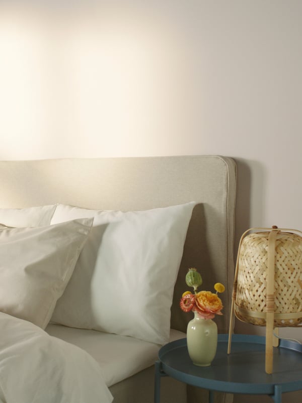 Lampe KNIXHULT en bambou et vase de fleurs sur une table d’appoint au coin d’un lit revêtu de textiles DVALA en coton blanc.