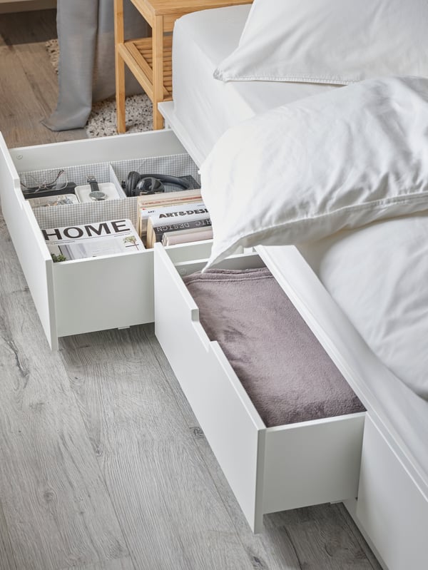 A NORDLI bed with storage with ÄNGSLILJA bed linen. Two drawers are open and contain books, a bedspread and other items.