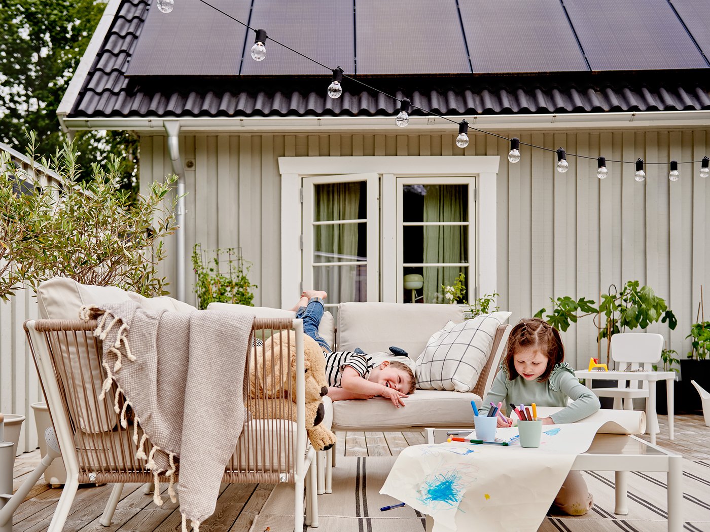 Zwei Kinder sind auf einer Terrasse. Eines liegt auf einem Sofa, während das andere an einem Tisch malt. Die Terrasse ist an ein Haus mit einem Solarpaneeldach angebaut.