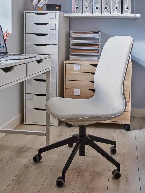 A beige conference chair with black legs and castors beside a white desk, and white and wooden drawer units.