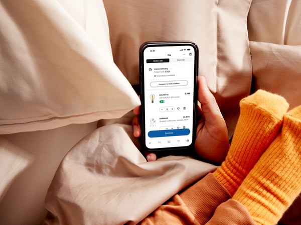 Na posteli drží někdo v ruce chytrý telefon s aplikací IKEA, na paži této osoby má položené nohy malé dítě v oranžových ponožkách.