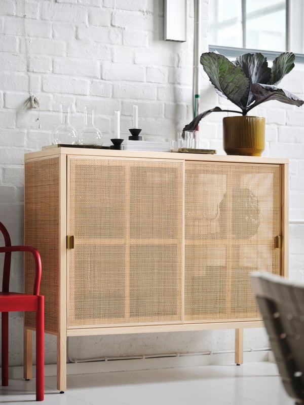 A red chair placed next to a STOCKHOLM 2025 cabinet with sliding doors with glasses and candlestick/tealight holders on top.