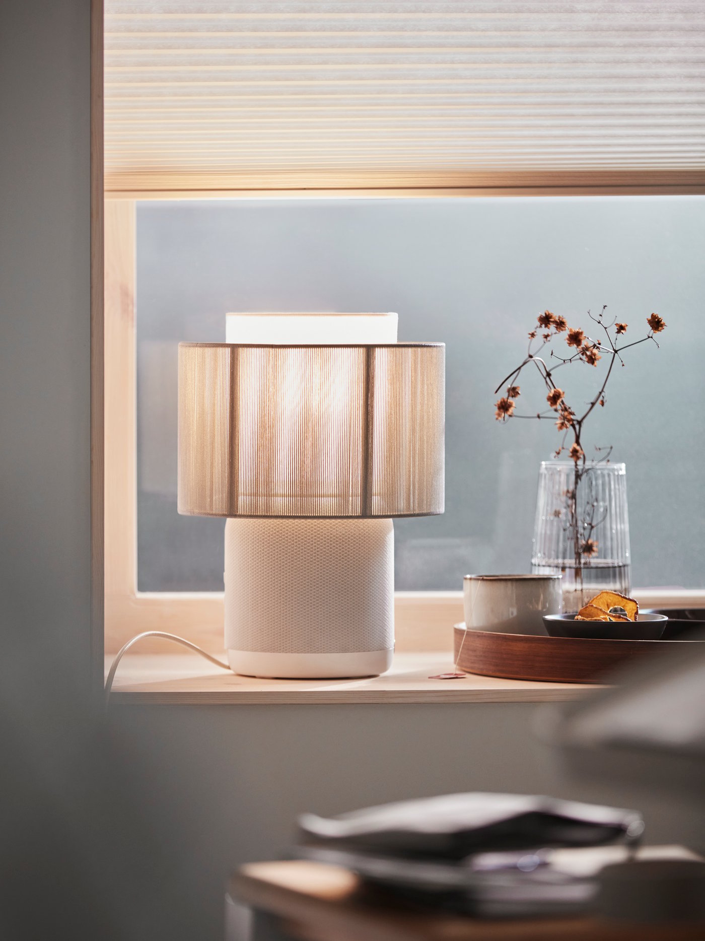 Vista de una ventana con una cortina roller IKEA. En el borde de la ventana hay una lámpara de mesa y una bandeja con accesorios decorativos.