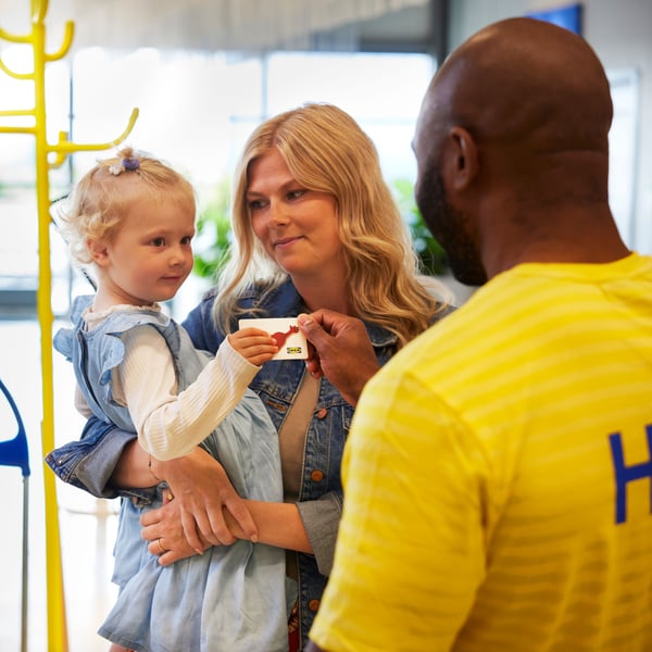 Malo dete dobija IKEA vrednosnu karticu od IKEA zaposlenog dok ga drži majka.