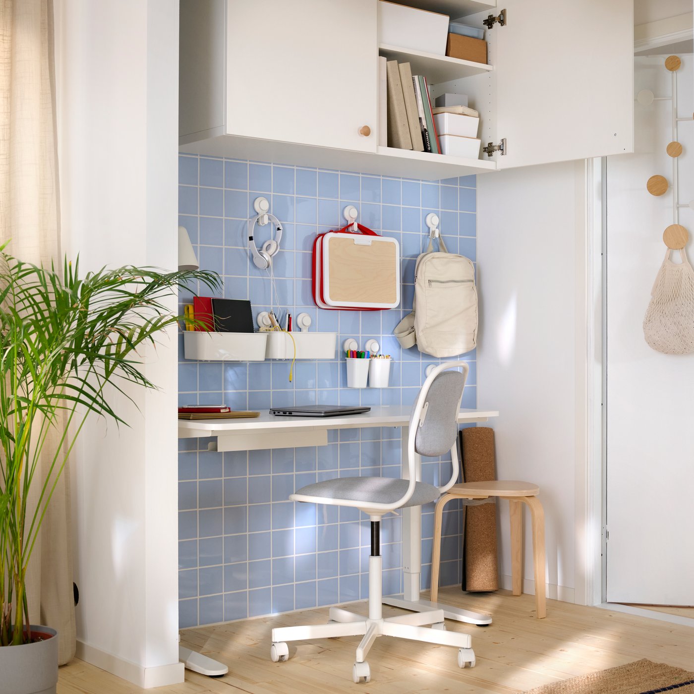 A creative corner with a light blue tile wall, a desk and work chairs. Lots of hooks on the wall and storage cabinets above.