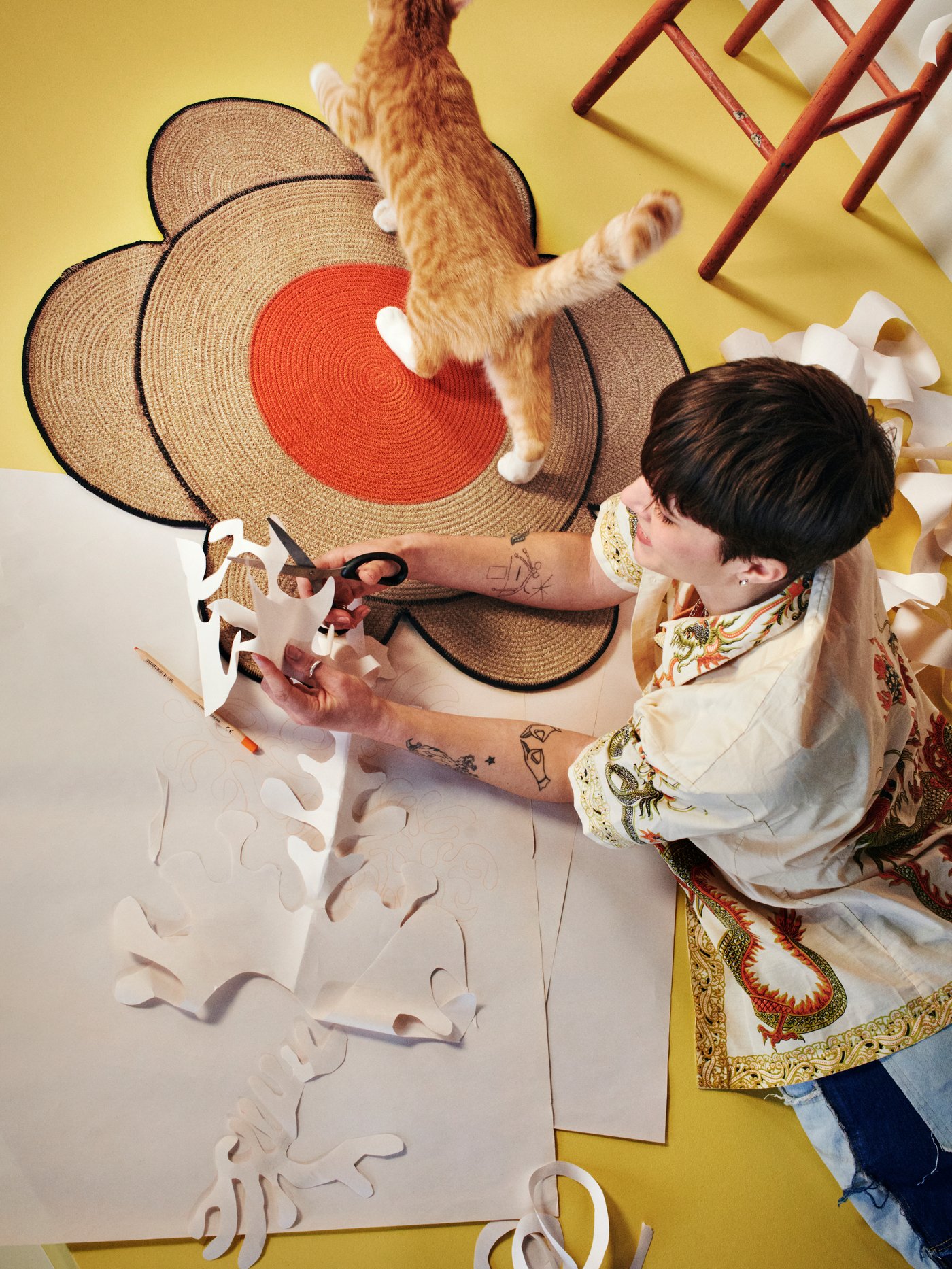A person lies on a yellow floor next to a cat and a flower-shaped natural MÄVINN rug.
