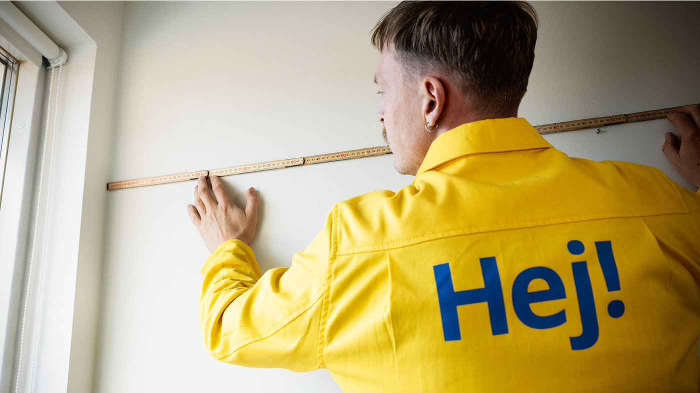 Person in yellow work jacket measuring a wall with a folding rule near a window.