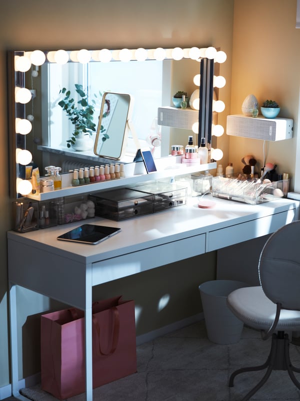 Un grand miroir rectangulaire avec des appliques LEDSJÖ en acier inoxydable tout autour, sur un bureau MICKE avec des organiseurs de maquillage.