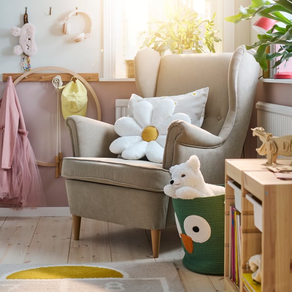 A beige STRANDMON wing chair in a bright children’s bedroom with a white flower-shaped SKOGSDUVA cushion on it.