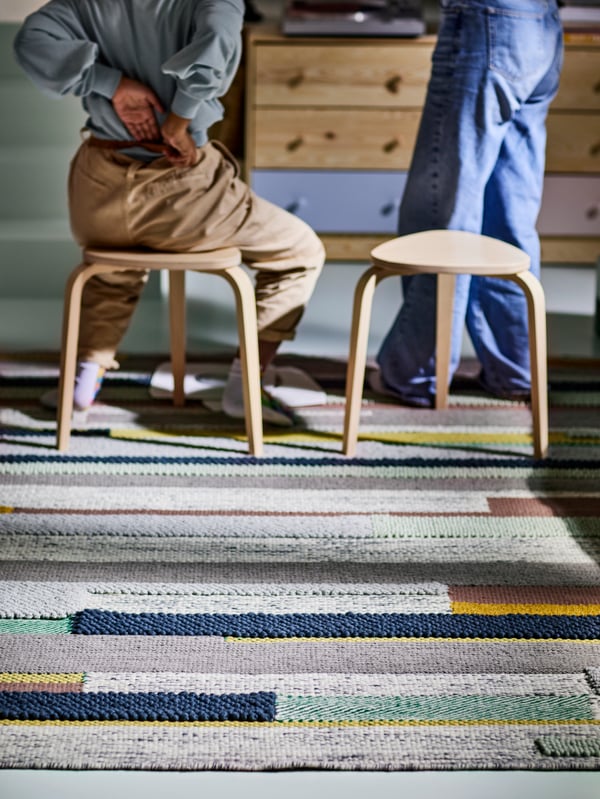 A multicolor BRÖNDEN rug with two birch stools on top, with one person standing and another sitting and storage behind.