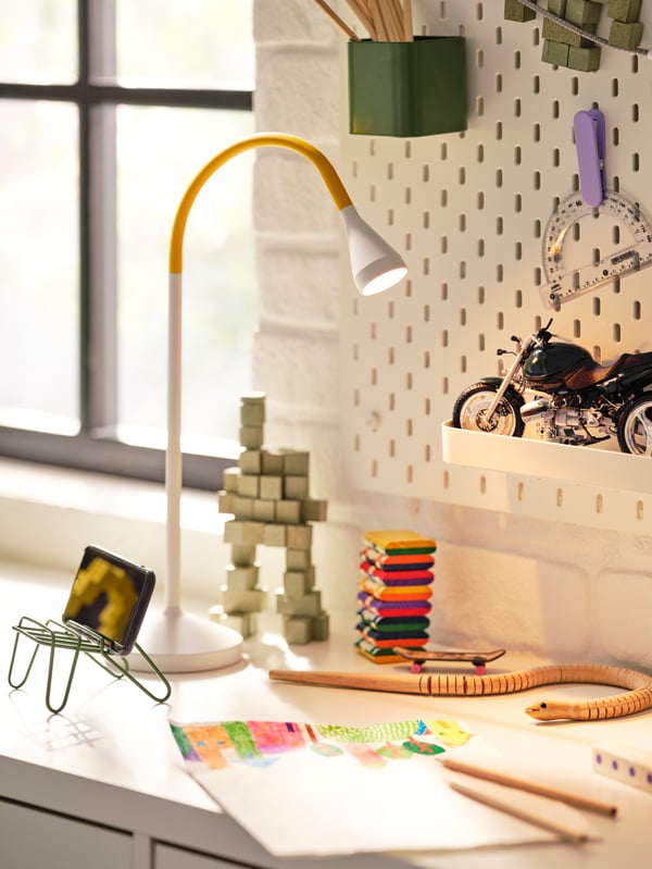 Close-up of drawing on white children’s school desk with green phone holder, white and yellow desk lamp and pegboard.