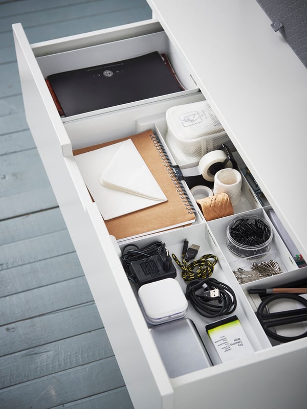 Un cajón de un escritorio blanco abierto, con una bandeja organizadora KUGGIS blanca en su interior. Dentro de los compartimentos se ven cables, artículos de escritorio y cuadernos.