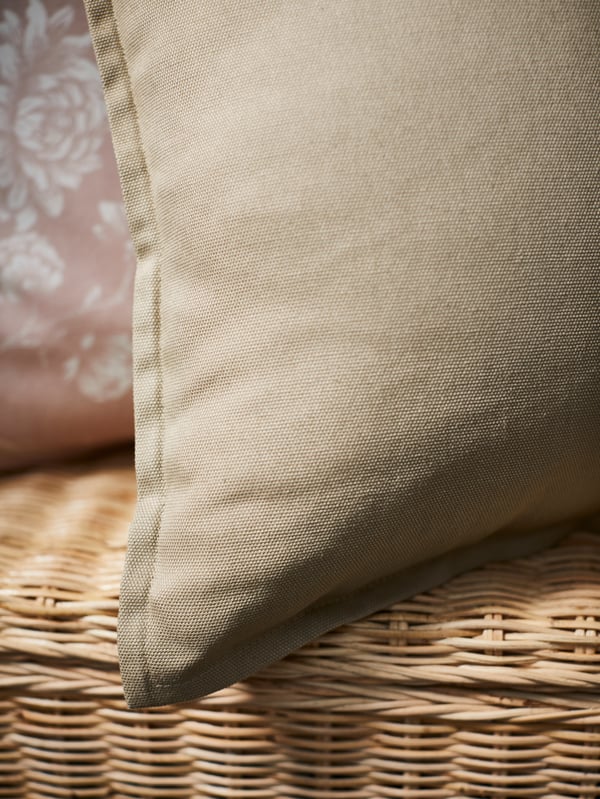 A corner of a cushion in a beige GURLI cover with a cushion in a light pink IDALINNEA cover behind it on a rattan bench.
