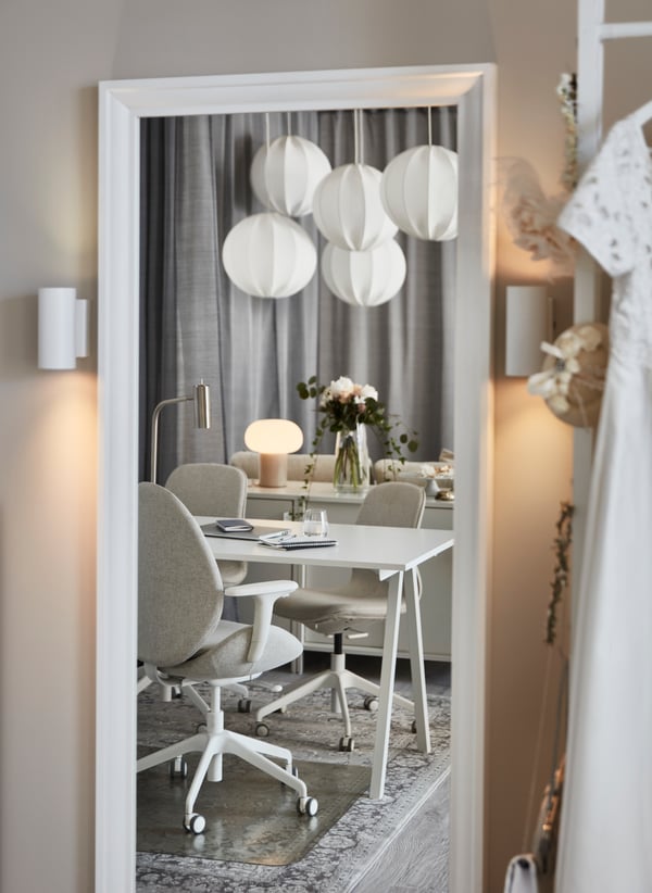 Su un grande specchio TOFTBYN con cornice bianca è riflessa l'immagine di uno spazio di lavoro luminoso e di un gruppo di lampadari a sospensione REGNSKUR bianchi.