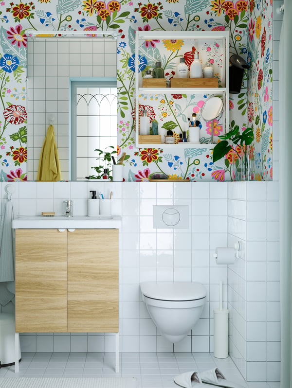 A washbasin, a mirror, open shelves above the toilet and floral wallpaper covering the upper half of the bathroom wall.