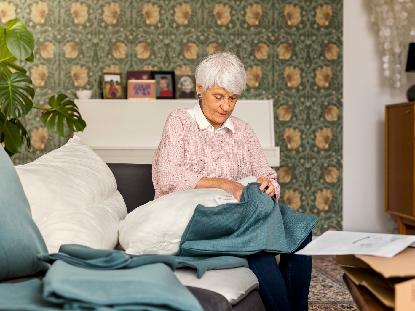 An elderly woman wearing a light pink sweater sits on a sofa while stuffing a sofa cushion in a new blue cover.